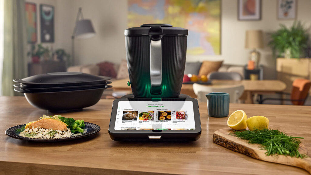 Thermomix TM7 kitchen appliance on wooden countertop surrounded by fresh ingredients and a plated salmon dish, with recipe touchscreen display visible.