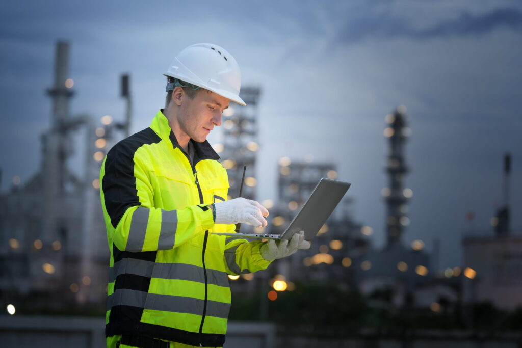 Worker in safety gear using laptop at industrial site during twilight reactive and predictive maintenance